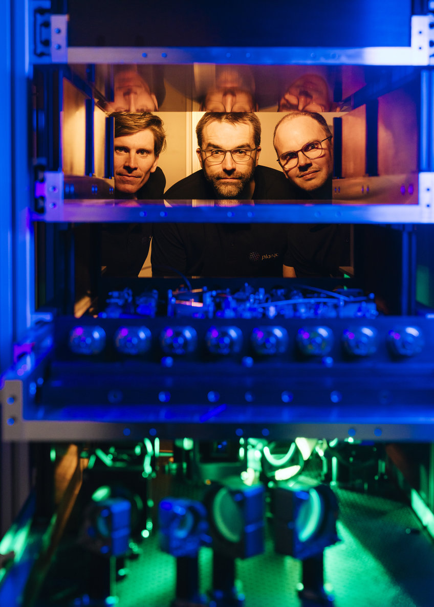Three men in a laboratory setting with complex machinery illuminated by blue and green lights, showcasing detailed reflective surfaces and various technical elements.