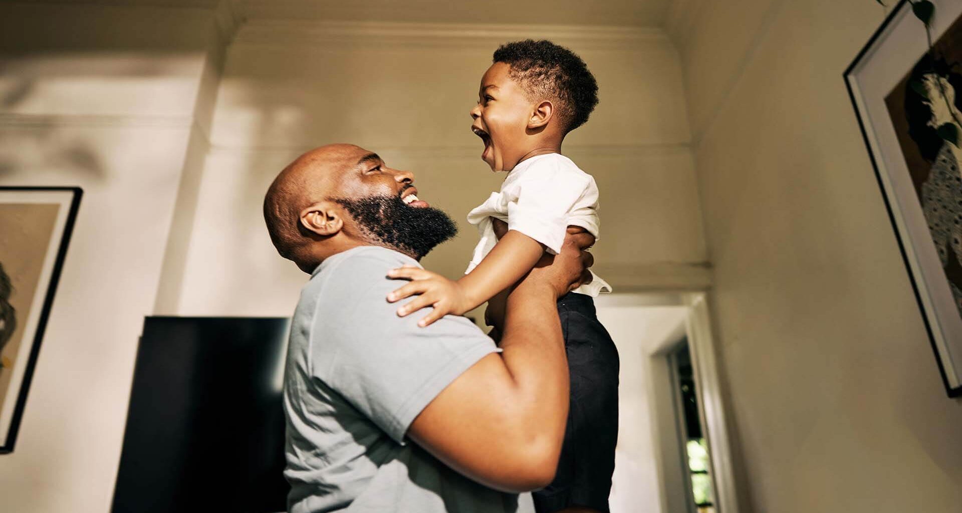 A non-custodial father celebrates his new job by lifting his toddler into the air of a sun-dappled room.