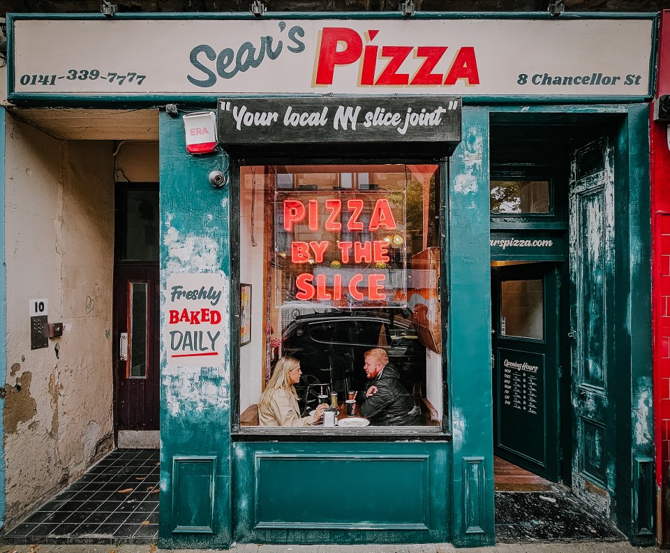 The exterior of Sear's Pizza in Glasgow, Scotland, with two customers visible through the window and a red neon sign that says "Pizza by the slice."