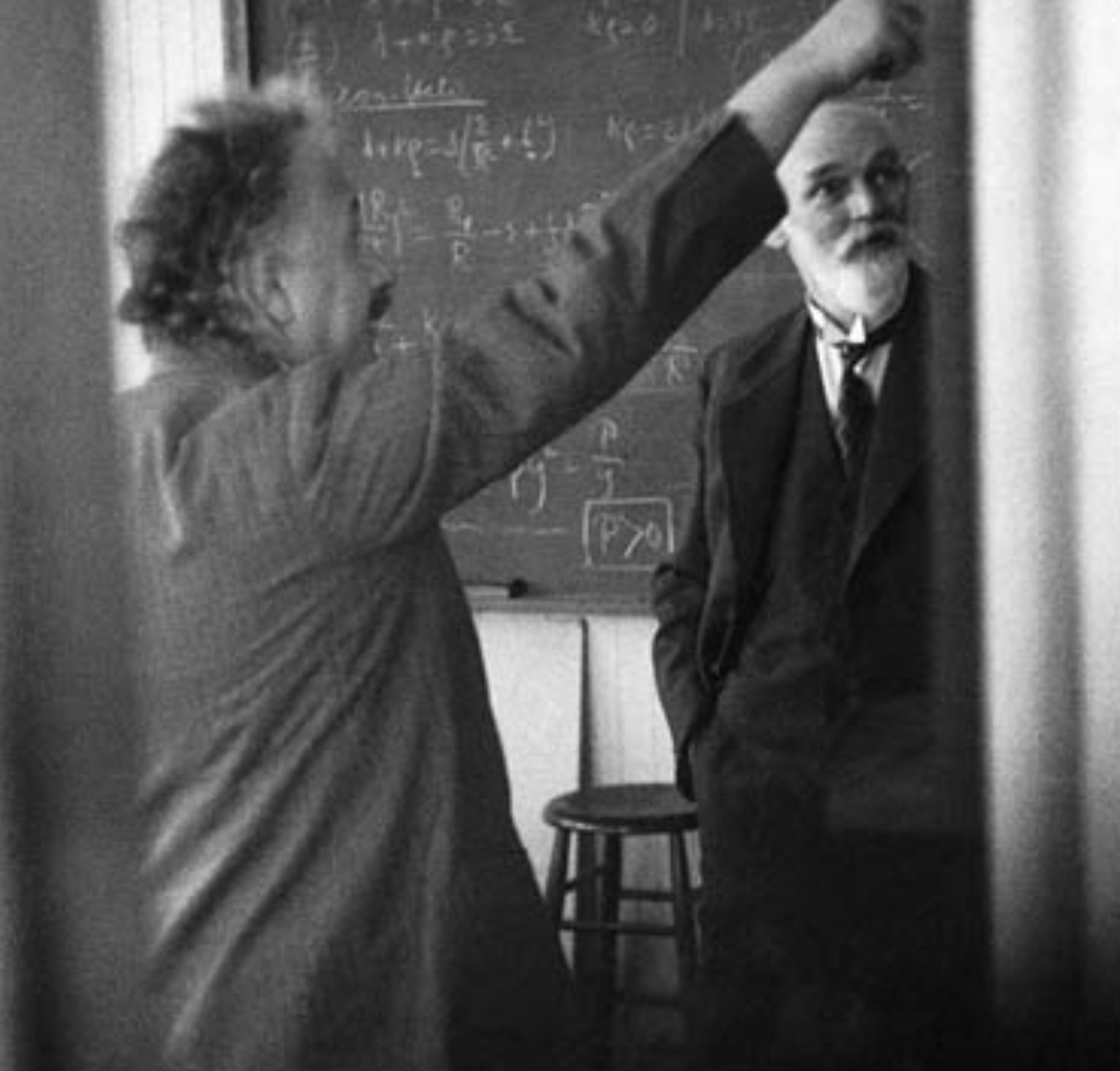 Black and white photo of two men in a classroom. One gestures towards a chalkboard filled with equations.