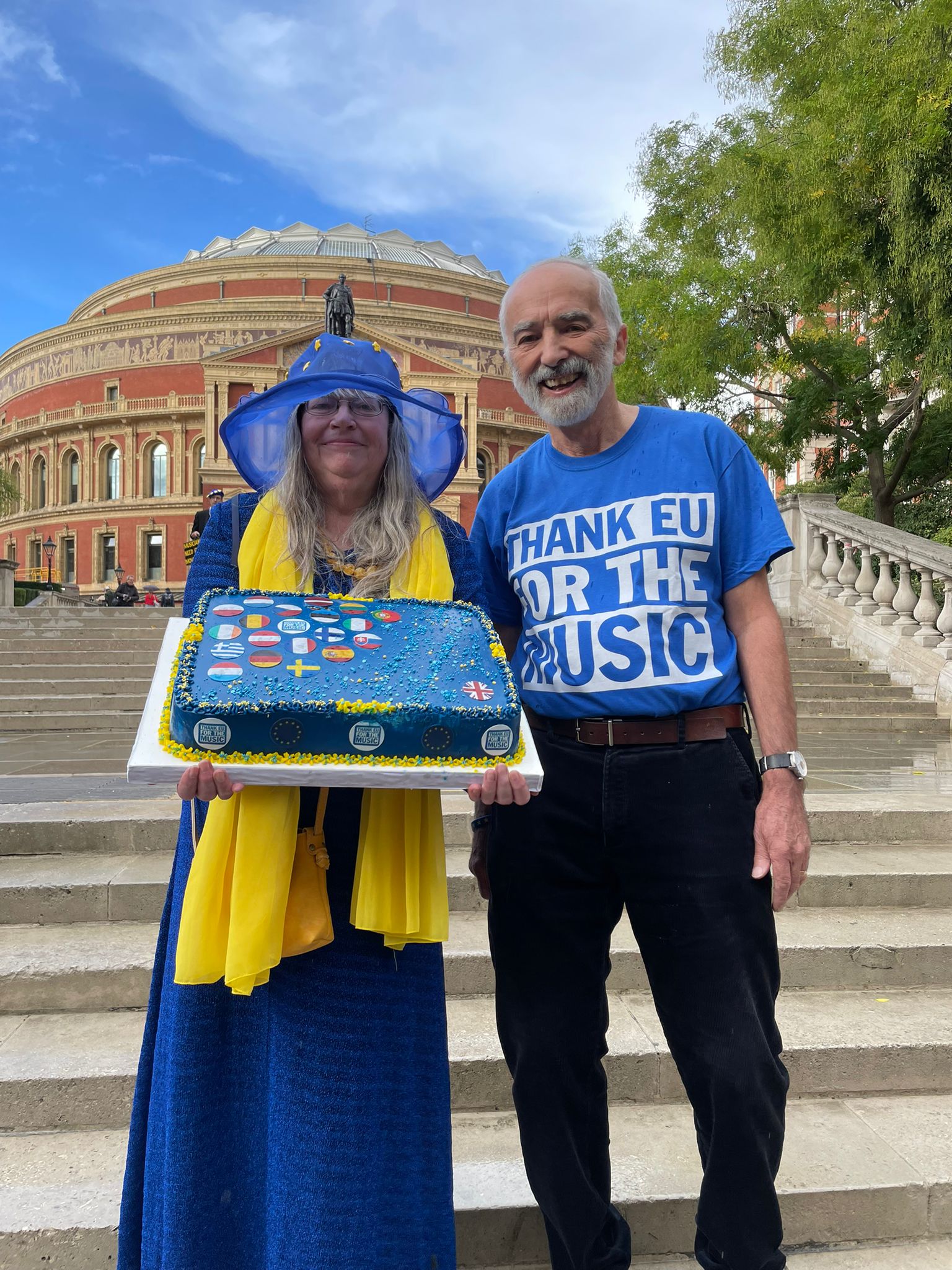 Two Thank EU for the Music campaigners stand outside the Royal Albert Hall. One is wearing a custom-made EU hat, with yellow scarf and blue dress, and holding a cake made up of the EU flags. 