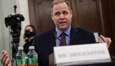 A man in a suit sits behind a table with open arms.