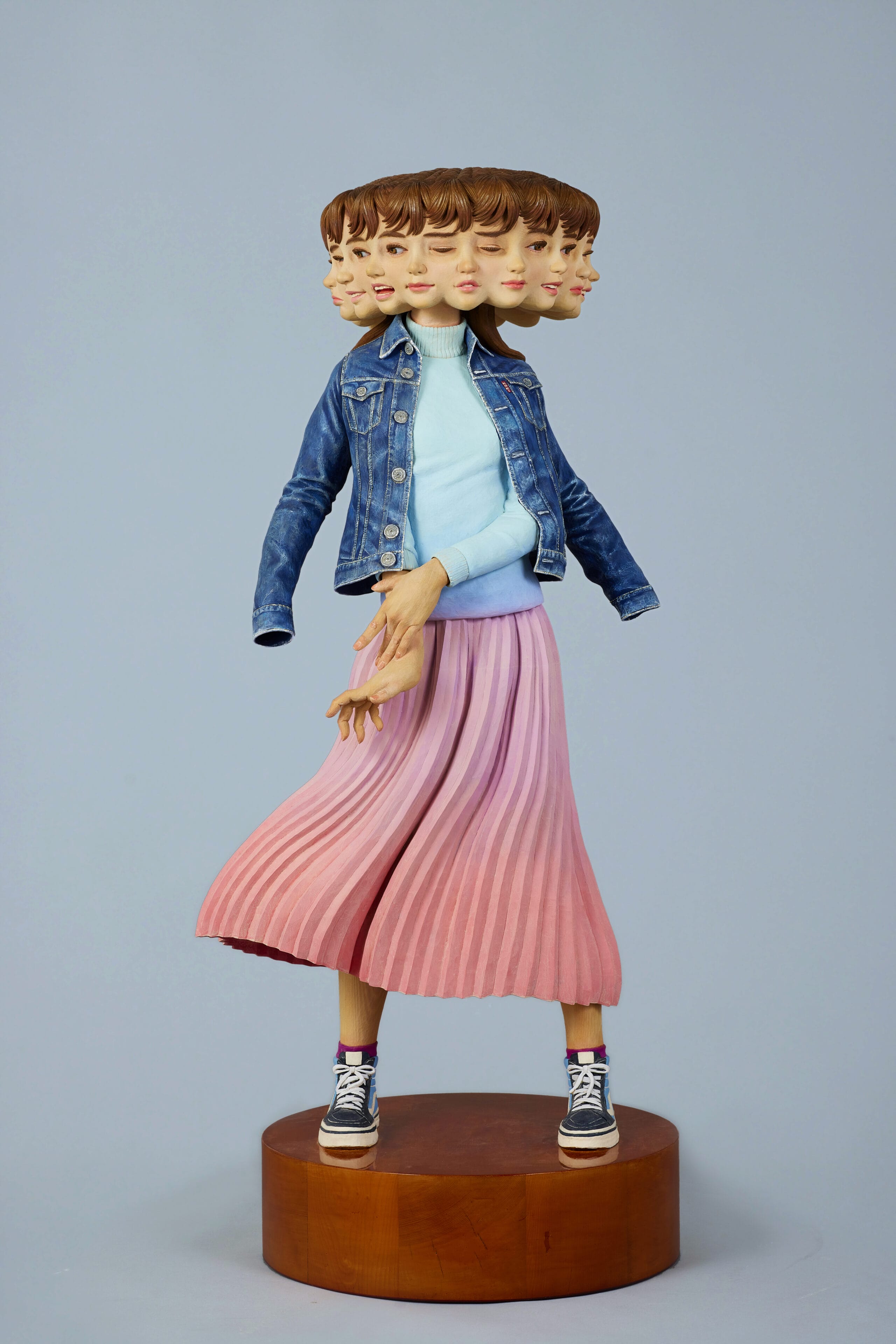 a wooden sculpture by Yoshitoshi Kanemaki of a female figure with a bright pink skirt and a denim jacket, with multiple faces and expressions