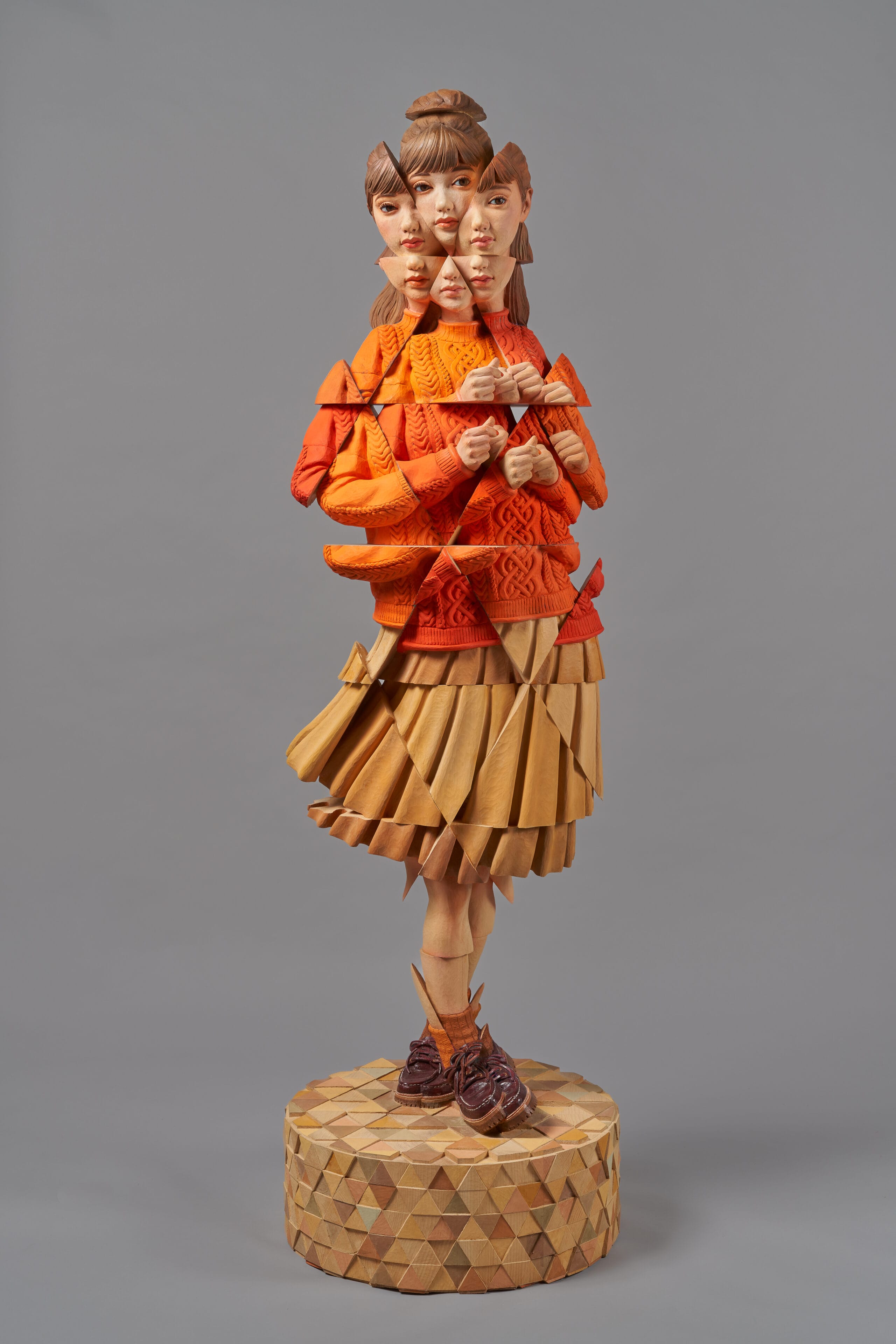 a wooden sculpture by Yoshitoshi Kanemaki of a female figure wearing an orange sweater and skirt, with prismatic or fragmented features