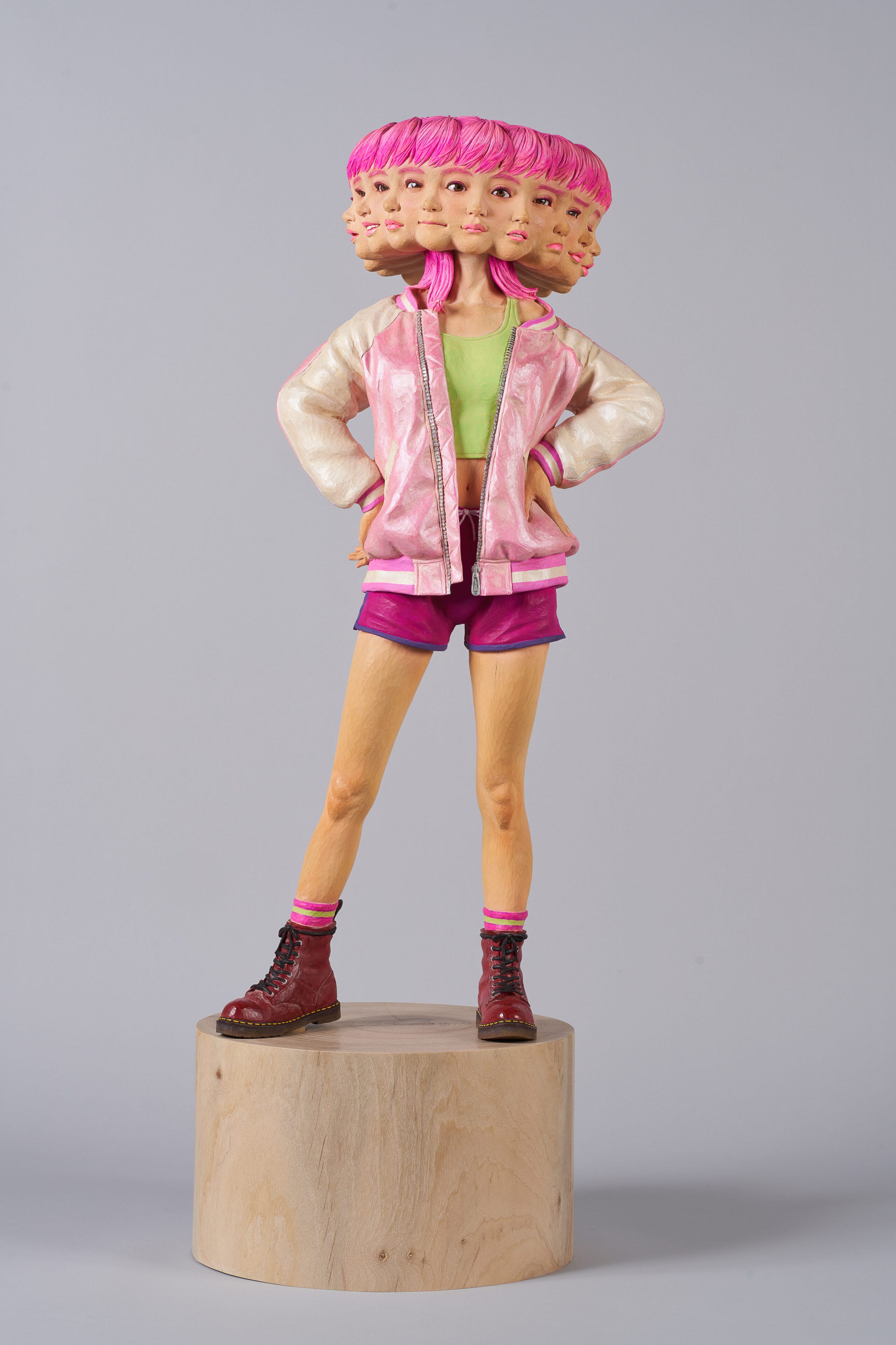 a wooden sculpture by Yoshitoshi Kanemaki of a female figure with bright pink shorts and a jacket, with multiple faces and expressions