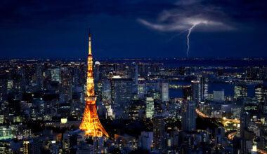 Tokyo skyline at night with a vibrant orange Tokyo Tower illuminated. A bolt of lightning strikes in the distant dark sky, creating a dramatic scene