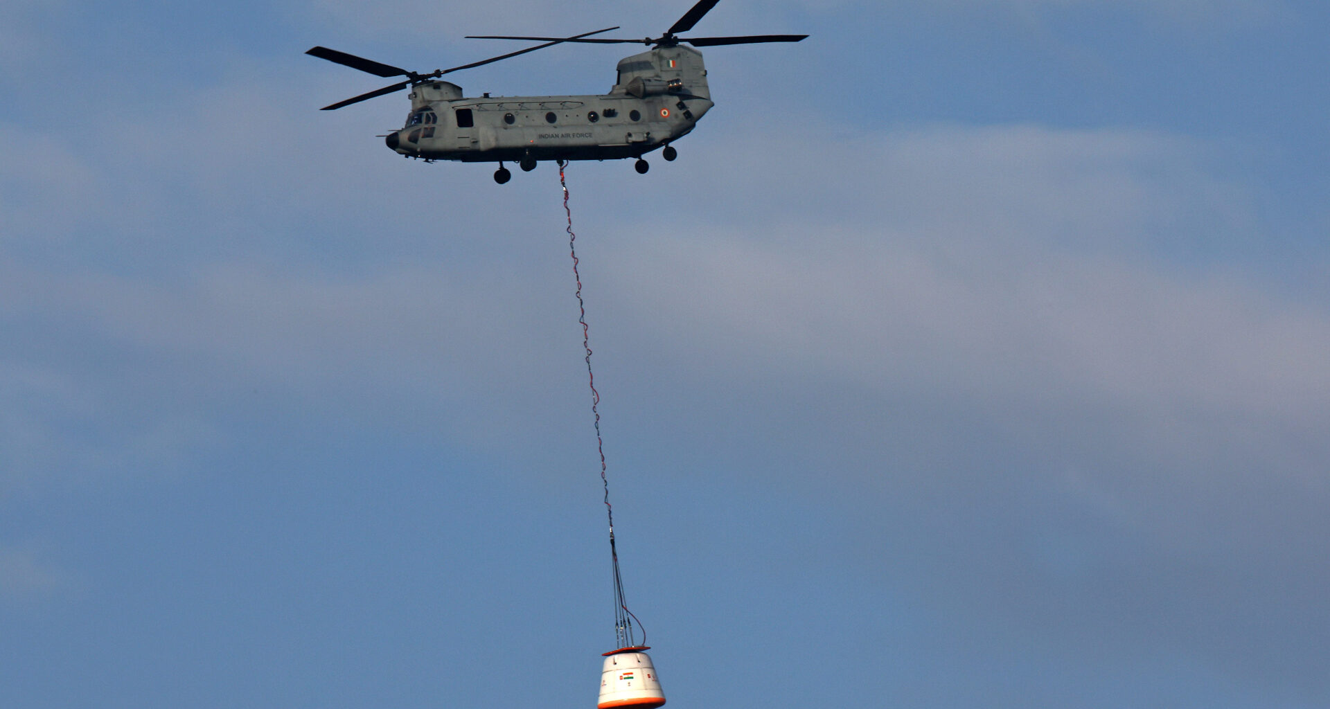 A helicopter lifts a large white crew module hoisted by a long rope in the air