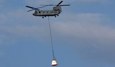 A helicopter lifts a large white crew module hoisted by a long rope in the air