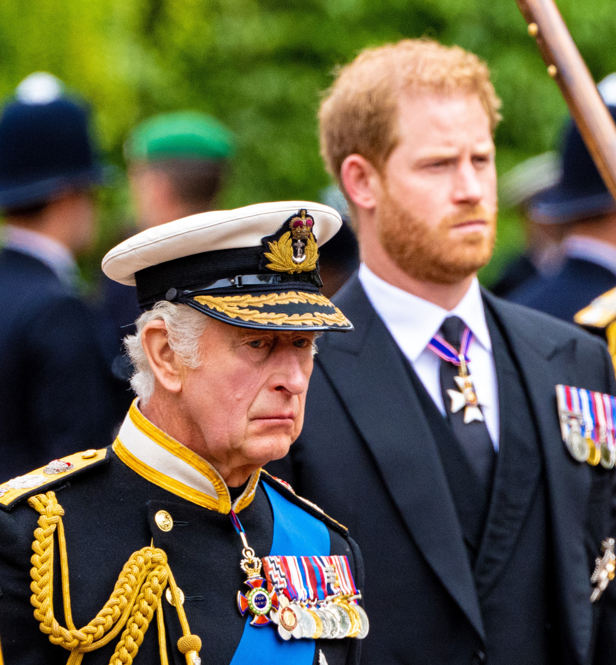 King Charles III in naval uniform with Prince Harry behind him in a suit.