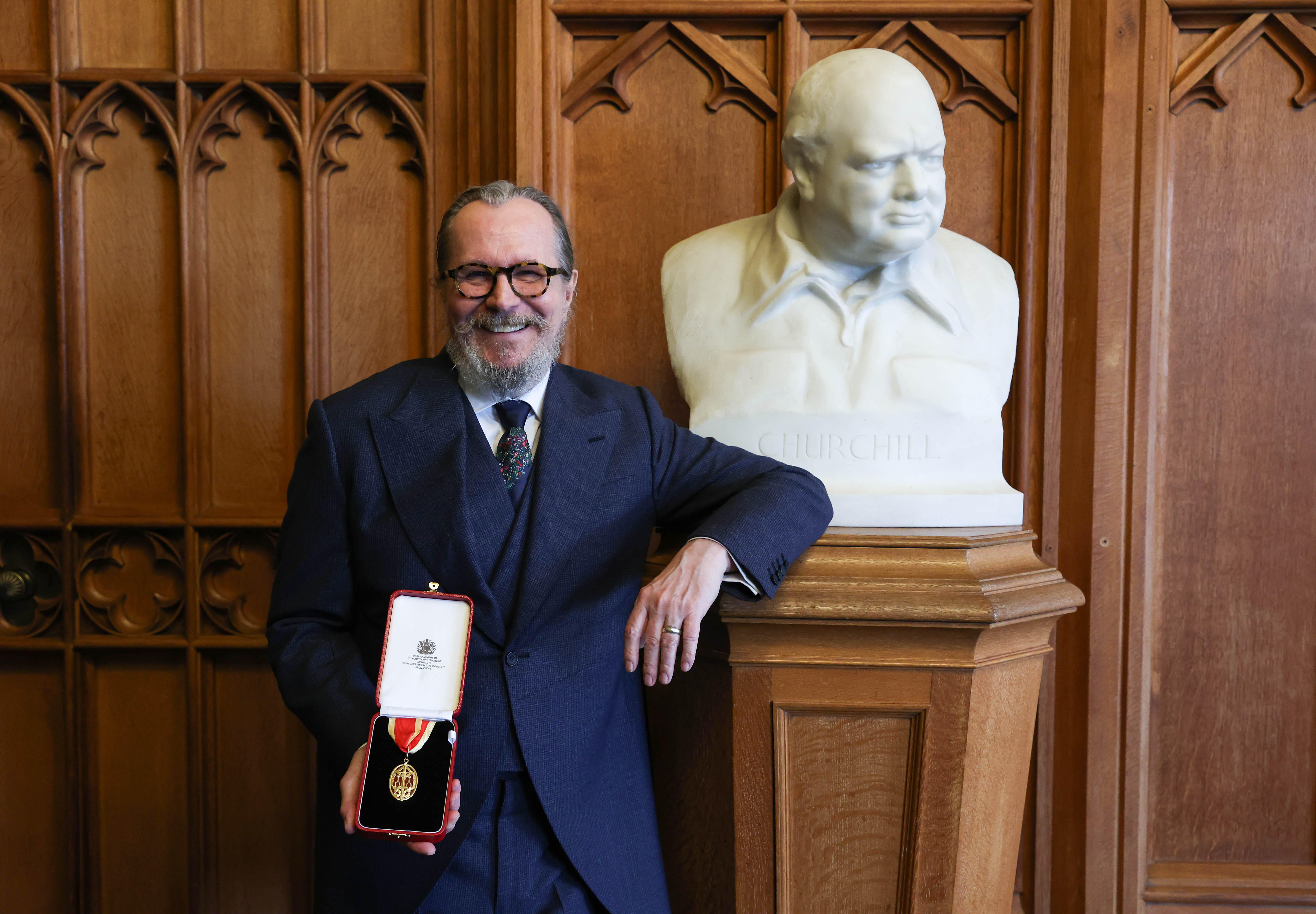 Sir Gary Oldman, smiling, in a dark suit, holding his knighthood medal in an open red case next to a bust of Winston Churchill.