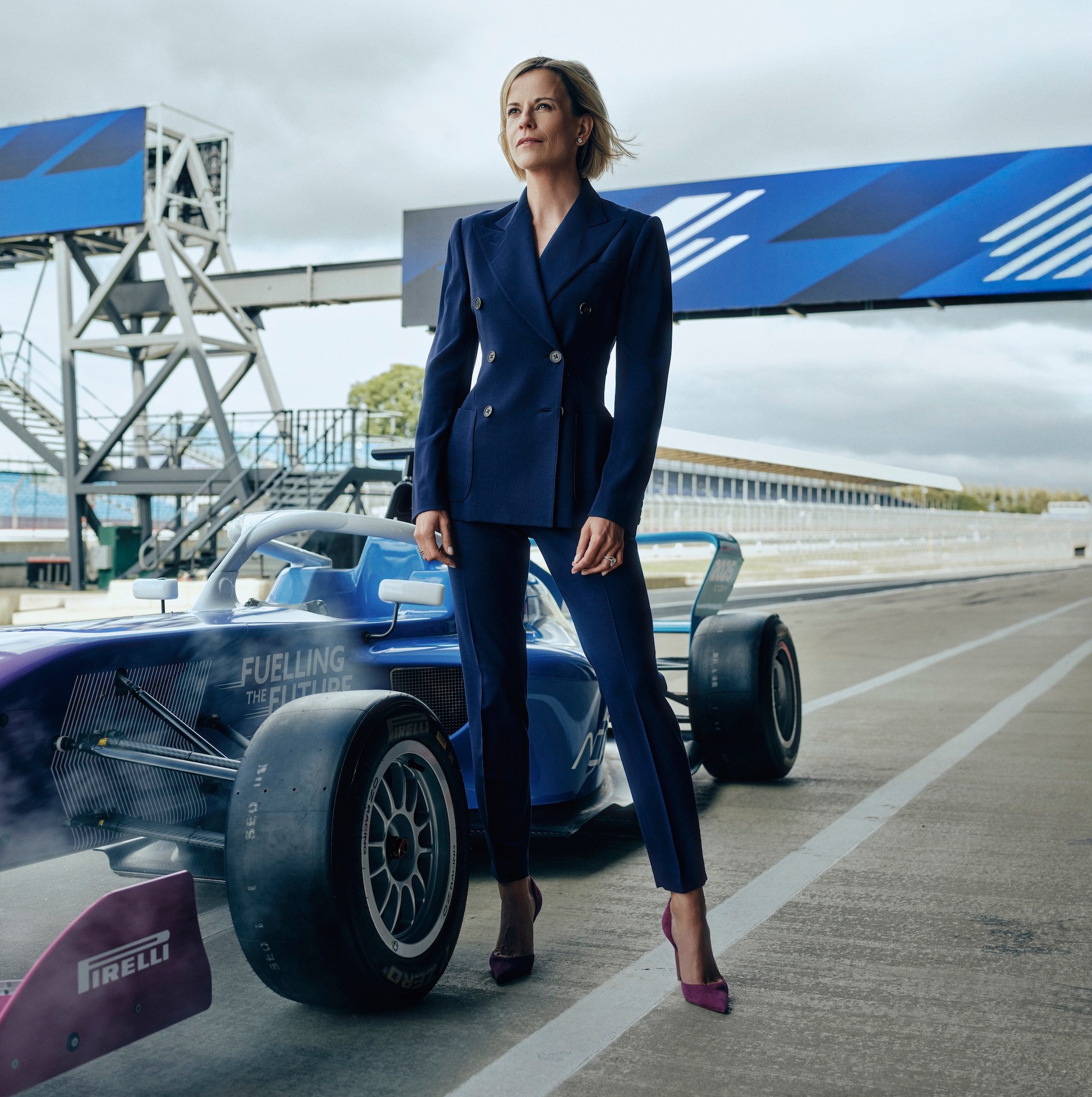 Susie Wolff stands in a dark blue suit next to a blue race car on a racetrack.