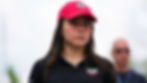 Woman in red cap and black shirt with TAG Heuer logos, looking determined. Blurred background, person in sunglasses behind her. Bright day.