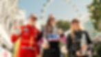 Three racing drivers in colorful suits and Pirelli caps hold trophies. Ferris wheel in the background; a joyful, victorious mood.