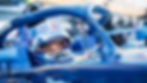 Race car driver in a blue vehicle, wearing a helmet, looks focused. Sunlit racetrack setting with blurred background.