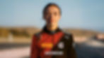 A woman in a red and black racing suit with logos stands on a racetrack, looking confident. The background is a blurred racetrack scene.