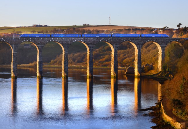 A view of a blue train riding over a river on a tall, arched bridge.