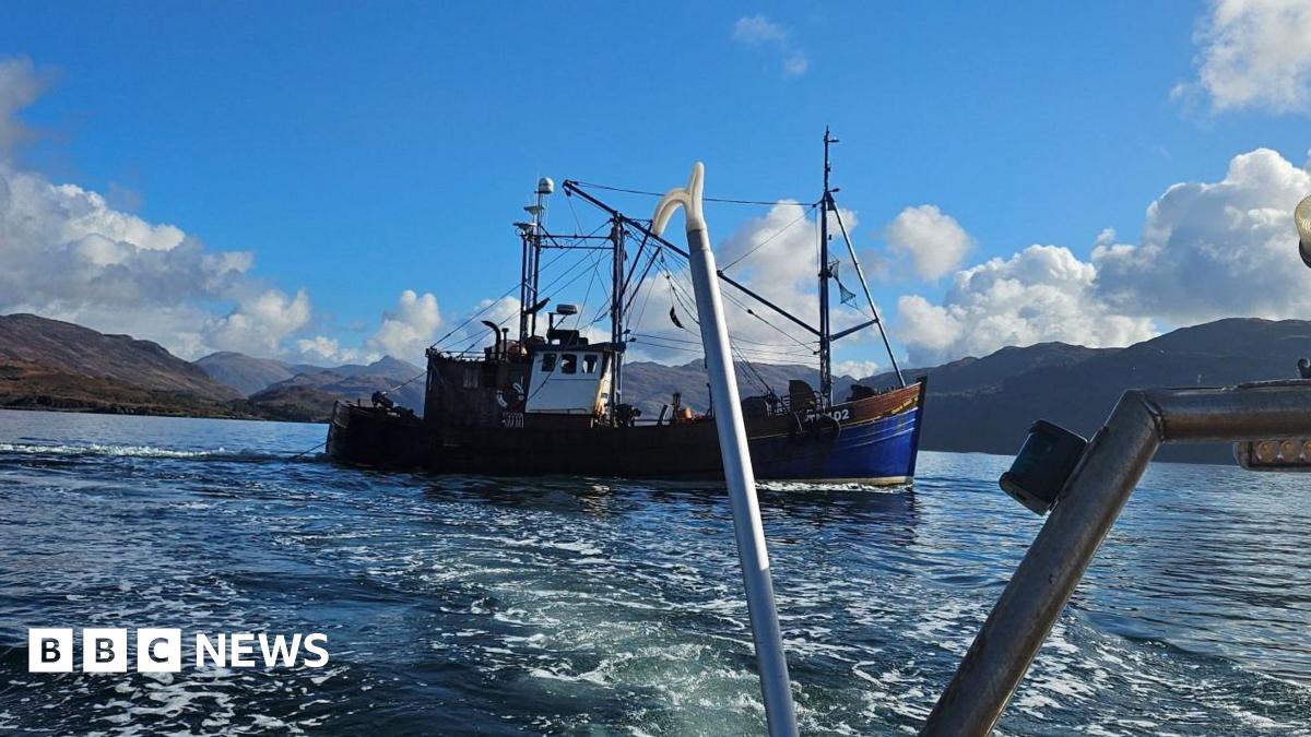A blue boat with red trim is seen passing through the water with mountains behind and a blue sky