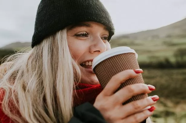 A beautiful, young woman is outdoors and holds a takeaway coffee cup and takes a drink. She is dressed in warm clothing to reflect the colder season. She smiles as she takes a sip of her warming beverage.