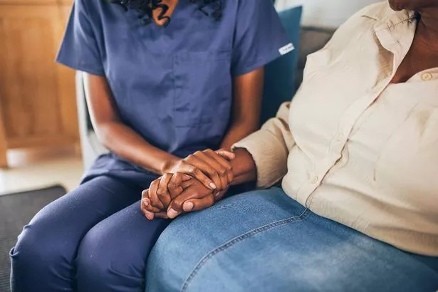 Close-up side view of in house nurse holding the hands of an elderly woman while sitting on a sofa at home. Young nurse comforting and reassuring senior woman