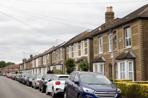 A view down a street where cars are parked on the road outside semi-detached houses with bay windows