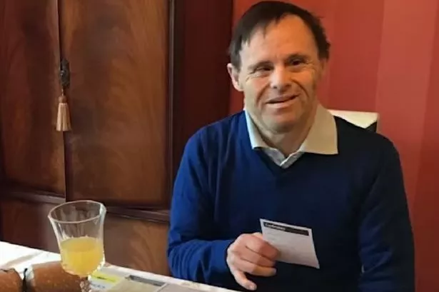 An image of a dark-haired man wearing a bright blue jumper, reading from a card.