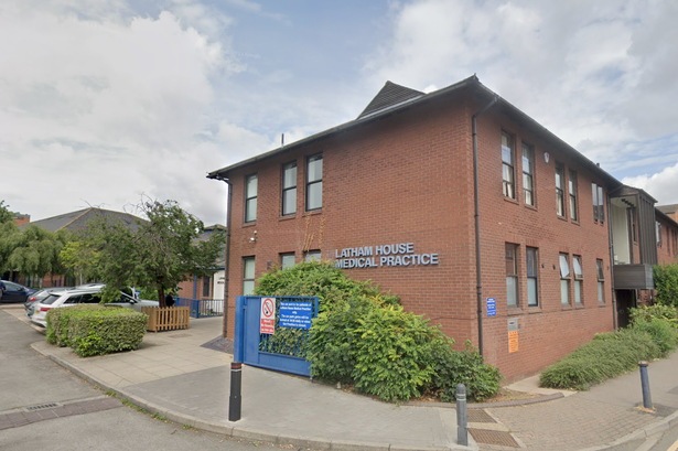 A brick building bears the sign Latham House Medical Practice. It is on the edge of a road, with bollards surrounding it.