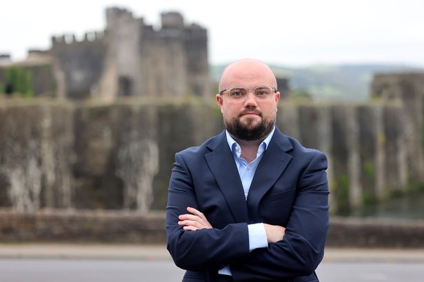 A man in a blue suit jacket with his arms folded in front of Caerphilly Castle