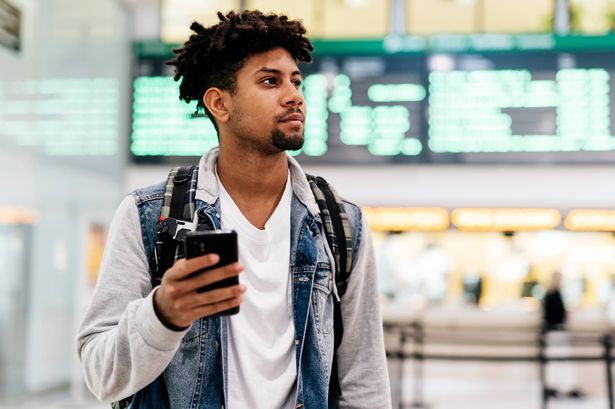 Young man inside the airport holding a mobile phone.young afro man holding a mobile phone looking at the departures of planes in an airport
