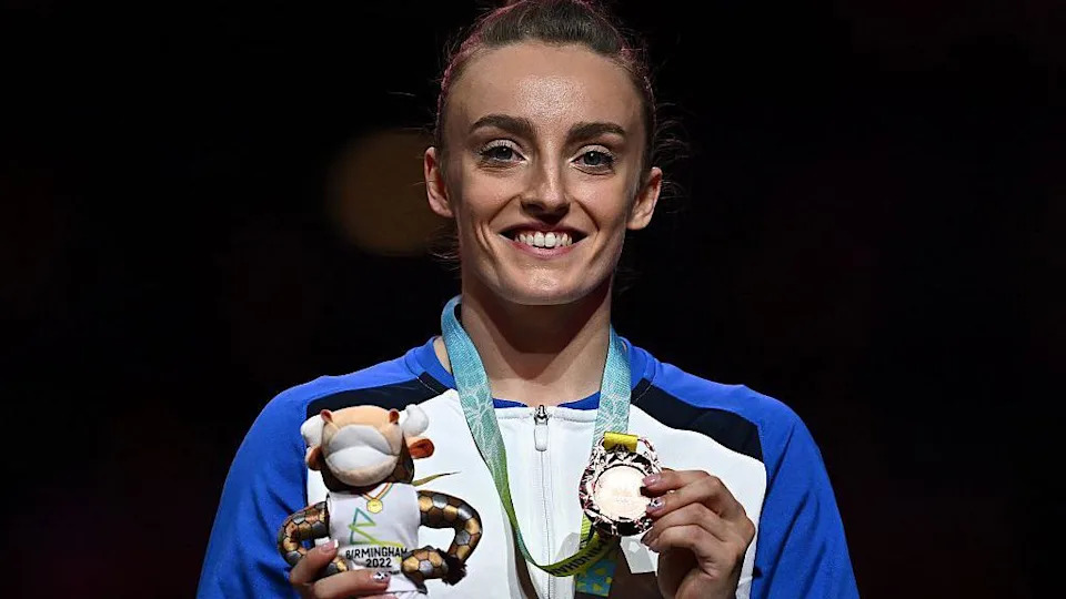 Gymnast Shannon Archer, a brown-haired woman with her hair in a tight bun, smiles at the camera as ahe holds up her bronze medal and a Birmingham bull mascot after being presented with her medal