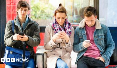 Three people sitting at a bus stop looking at their mobile phones