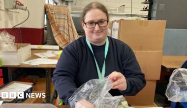 A woman is smiling at the camera. She is at work and looks like she's packaging something into a plastic bag. There are cardboard boxes behind her.