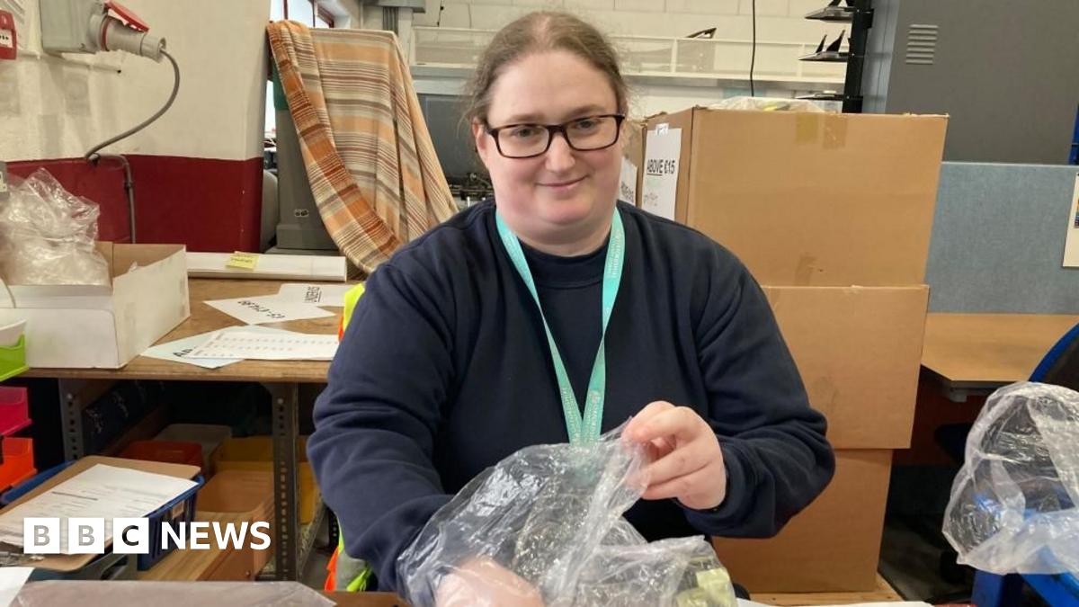 A woman is smiling at the camera. She is at work and looks like she's packaging something into a plastic bag. There are cardboard boxes behind her.