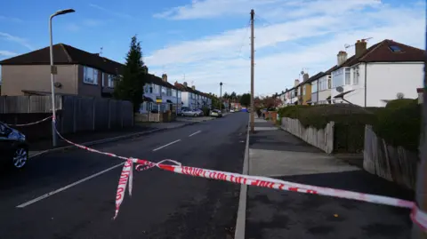 George Tryfonidis/BBC Red and white police tape is tied on lampposts across a road which has houses running alongside either side of it