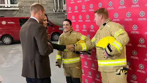 BBC A male firefighter with short ginger hair shakes hands with Catherine, Princess of Wales while a young female firefighter with dark hair stands beside them, smiling.  Prince William is standing beside his wife, talking to the recruits.  The firefighers are wearing beige safety suits and carrying bright yellow safety helmets. There is a red fire safety vehicle parked behind them.
