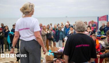 Ruth Hitchcock and Dave Hitchcock with their backs to the camera and facing a crowd of people. They are wearing t-shirts which say #resilientdefiance on the back in pink letters. The sky is grey. The sea is a light grey.