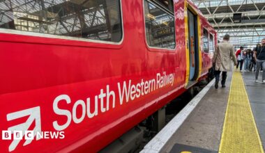 The side of a South Western Railways train in its red and white livery halted at a station, with the doors open.