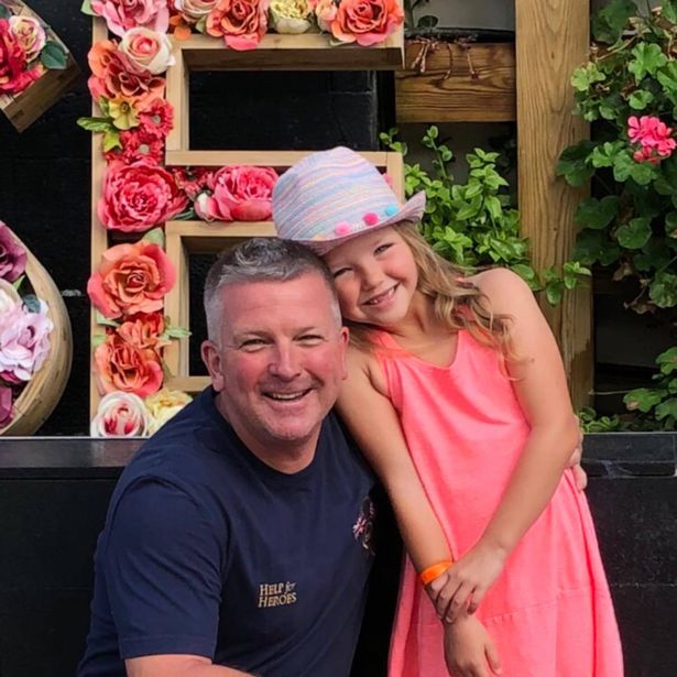 Jason sitting next to his daughter who is standing wearing a pink dress. 