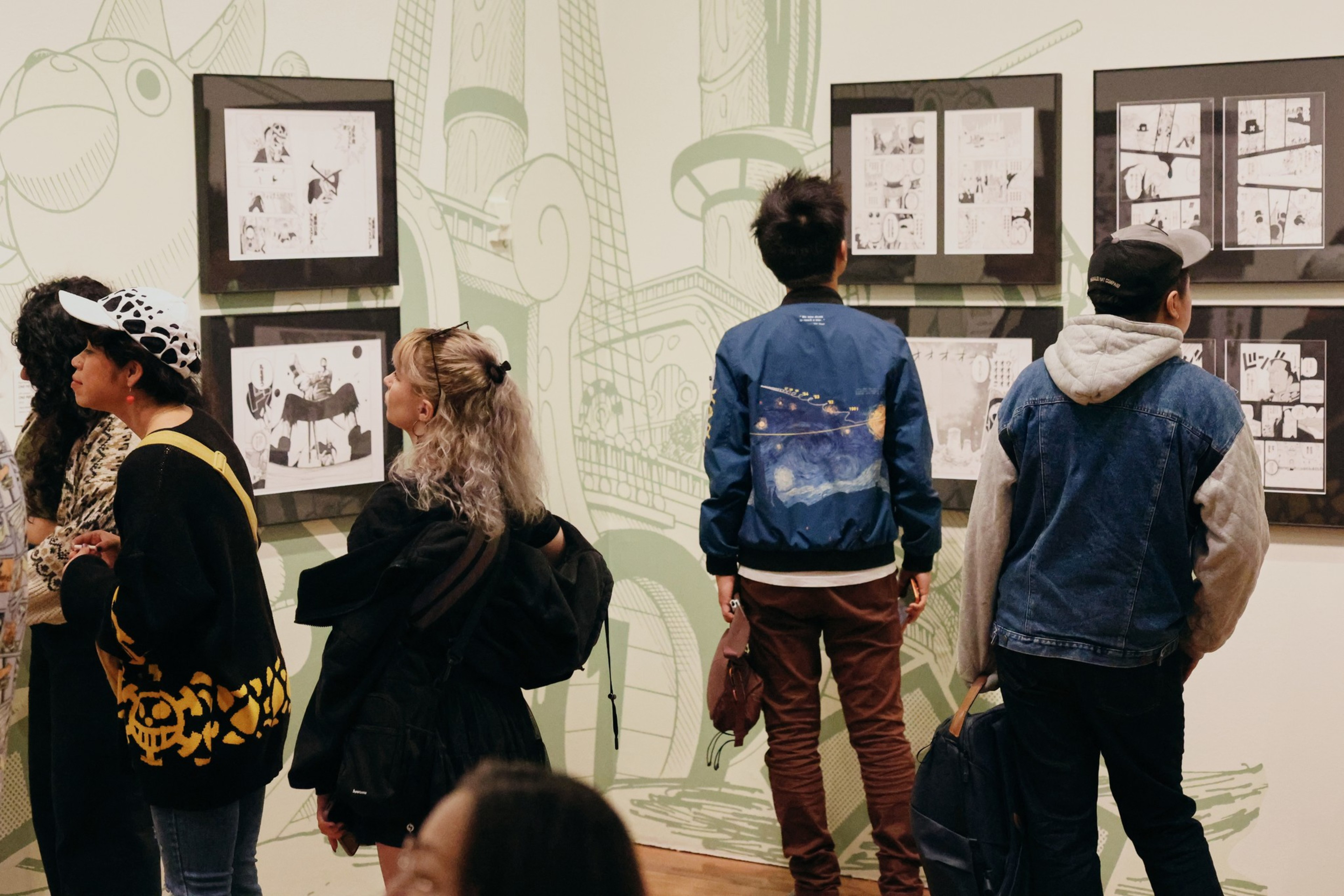 Five people stand closely, viewing framed black-and-white comic or manga panels displayed on pale green walls with large, abstract illustrations in the background.