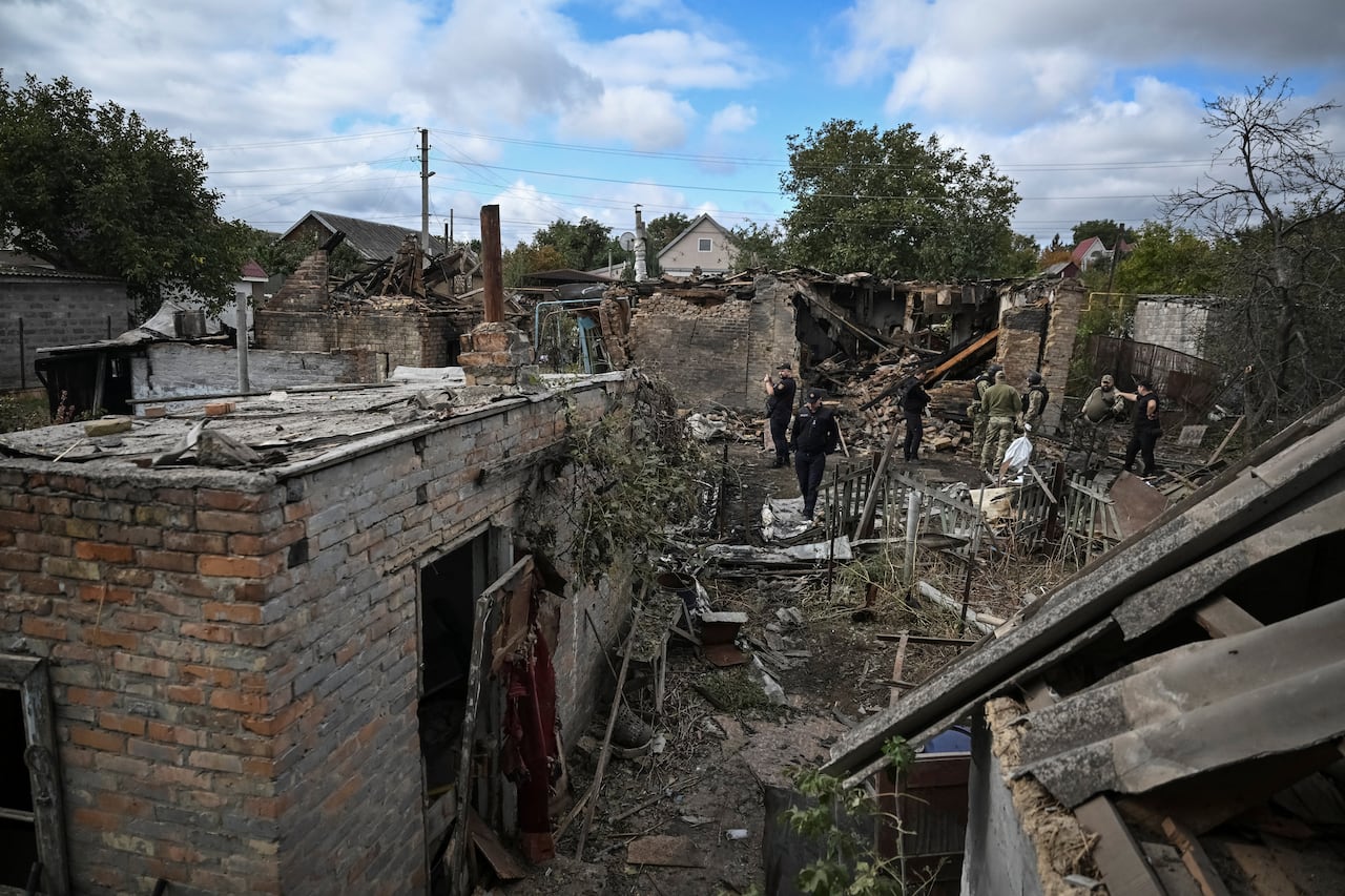 People are shown walking in between heavily damaged buildings, with debris strewn on the ground.