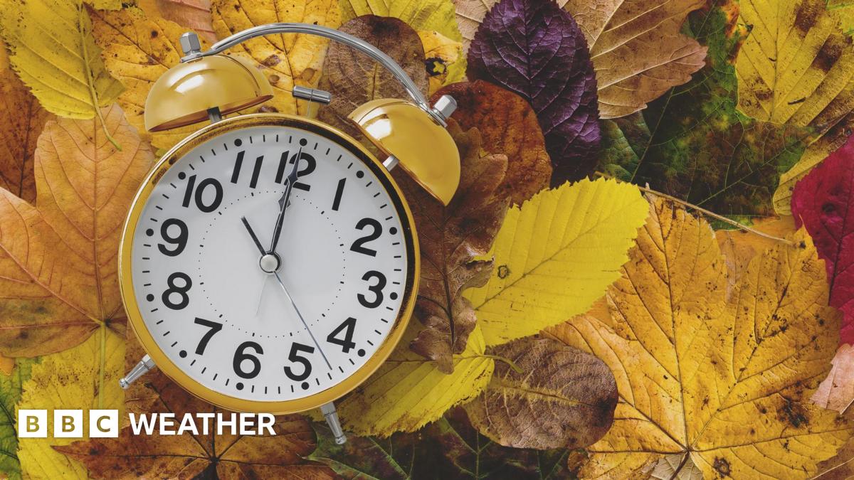 A stock photo of an old-fashioned analogue yellow alarm clock sitting on a pile of yellow, brown, green and red fallen leaves