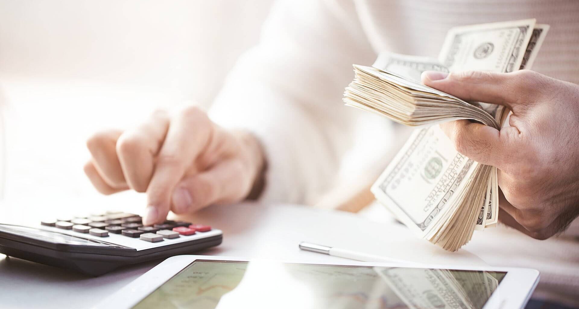 A close-up of two hands, one using a calculator and the other holding a stack of money.
