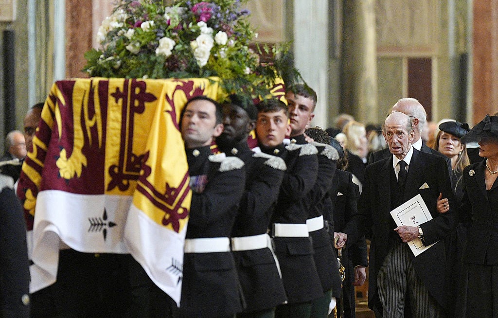 People walk behind a casket carried by military pallbearers and covered with a flag and a large floral arrangement.
