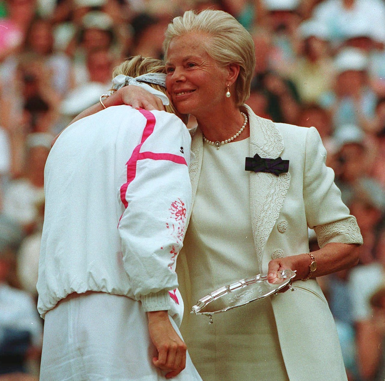 A person holding a silver plate comforts another person while standing on a tennis court.