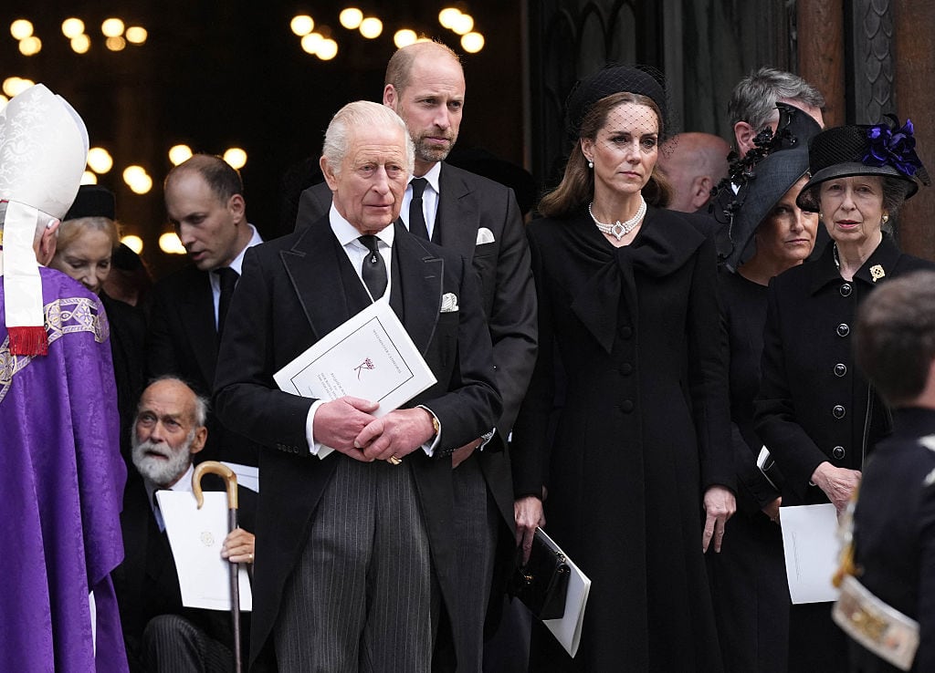 Several people wearing black stand in the entrance of a cathedral holding programs from a funeral.