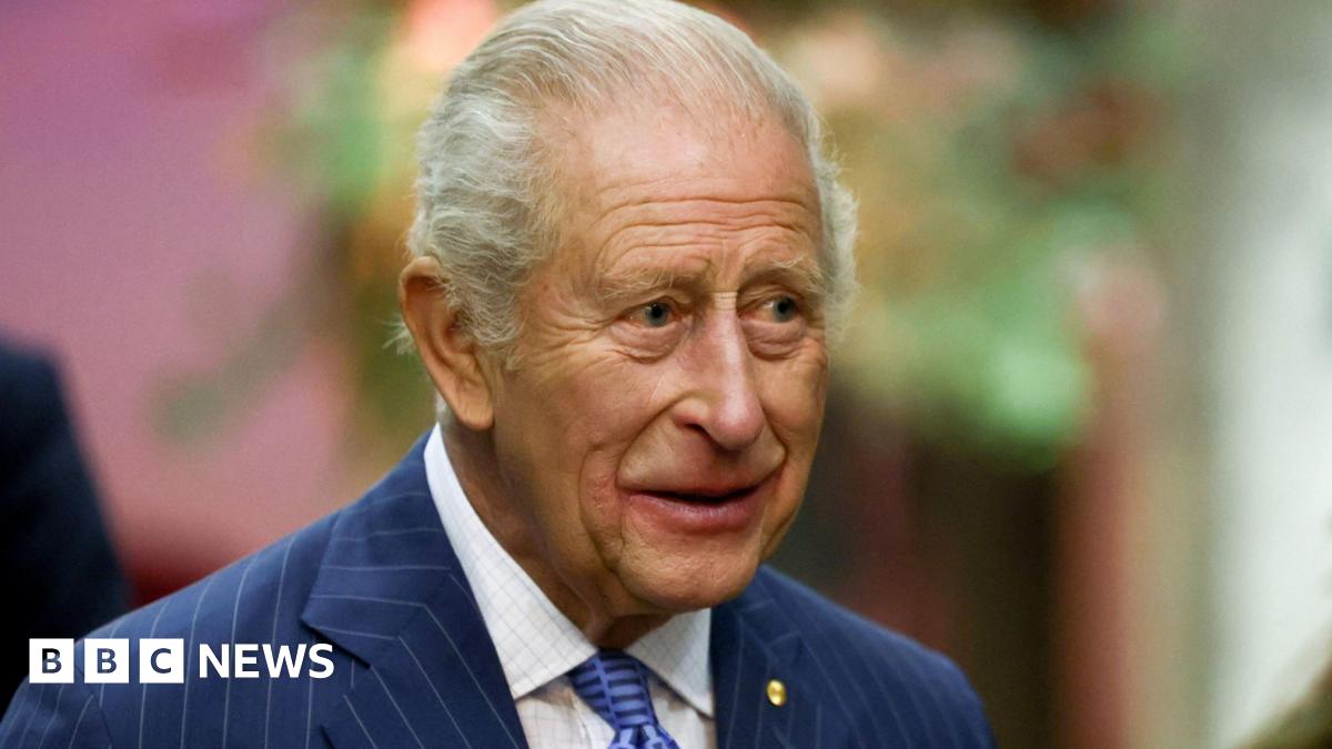 King Charles - an elderly man with short white hair - wearing a blue suit jacket with a gold pin on the left side, a blue tie and a white shirt, smiles as he looks to our right.