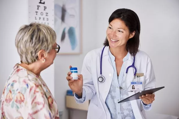 Smiling female medical professional explaining prescription medicine to patient. She is wearing lab coat and holding tablet PC. They are sitting in examination room.
