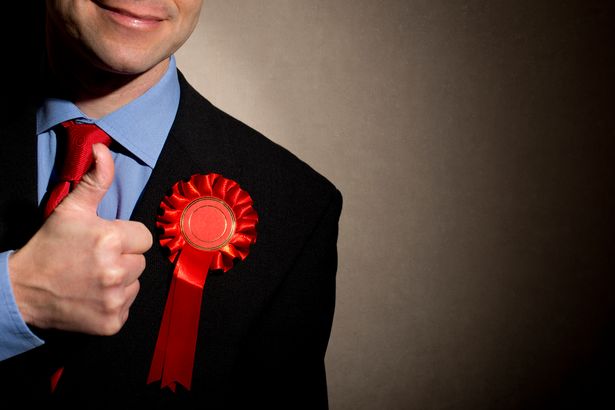 Smiling election candidate with a thumbs up hand gesture. The man is wearing a dark business suit with a red rosette