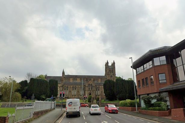 As road leading up to an intersection, in front of building. Cars, and vans seen travelling on the road
