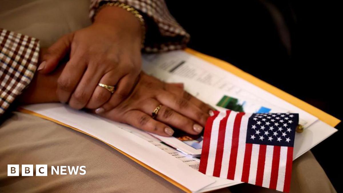A black woman's hands, with pink manicured nails and a two rings, are crossed i her lap on top of a small stack of papers and a small US flag