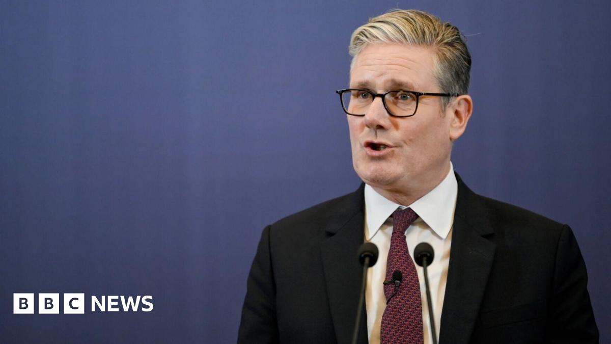 Sir Keir Starmer, dressed in a dark suit, white shirt, and red patterned tie, stands at a podium speaking during a news conference in Mumbai. Two microphones are positioned in front of him, and the backdrop is a plain blue screen.
